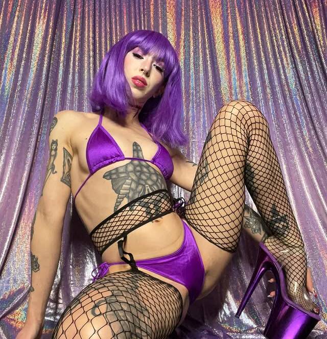 Person with a bright purple wig wearing a matching purple bikini and fishnet stockings posing against a shimmering backdrop with a high heel visible