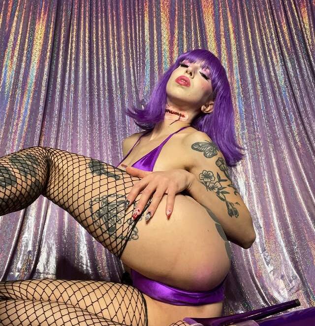 Person with bright purple hair and tattoos poses in a shiny purple bikini and fishnet stockings against a glittery backdrop leg raised toward the camera