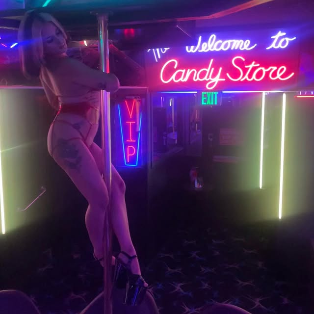 Woman pole dancing in a neon lit club with a bright Candy Store sign in the background