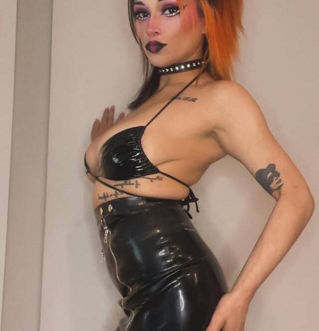Person in a black latex outfit posing sideways with bold makeup and orangeblack hair visible tattoos on arm and torso