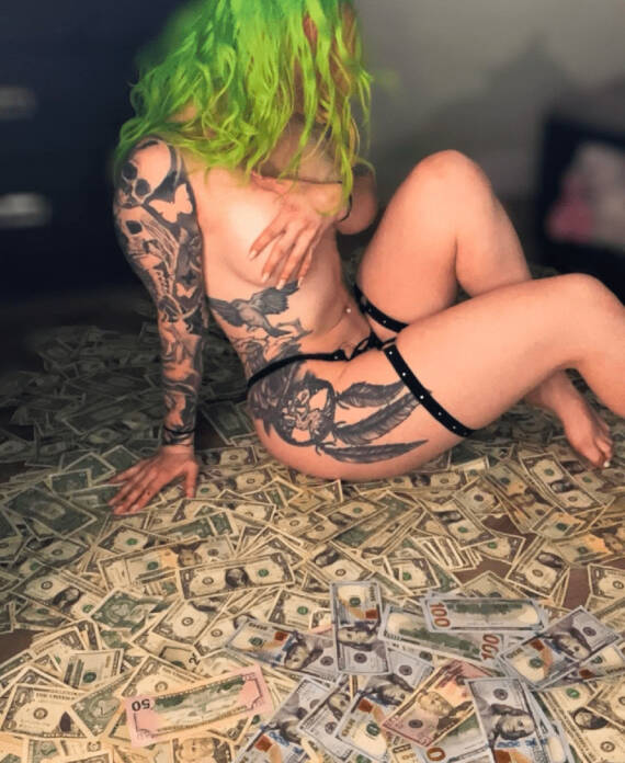 Person with neon green hair and full arm tattoos sits on a floor covered with scattered cash wearing black straps