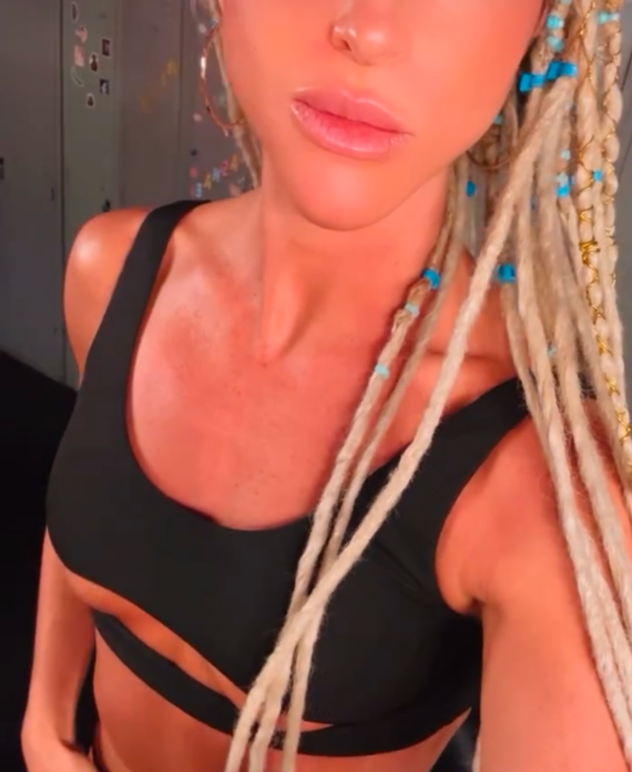 Close up selfie of a woman with long blonde dreadlocks wearing a black bikini top turquoise bead accents in her hair looking at the camera with blue eyes lockers visible in the background