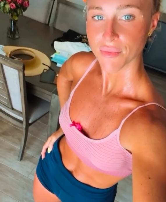 Woman in a pink sports bra and navy shorts taking a selfie in a bright kitchen with flowers on the table behind her