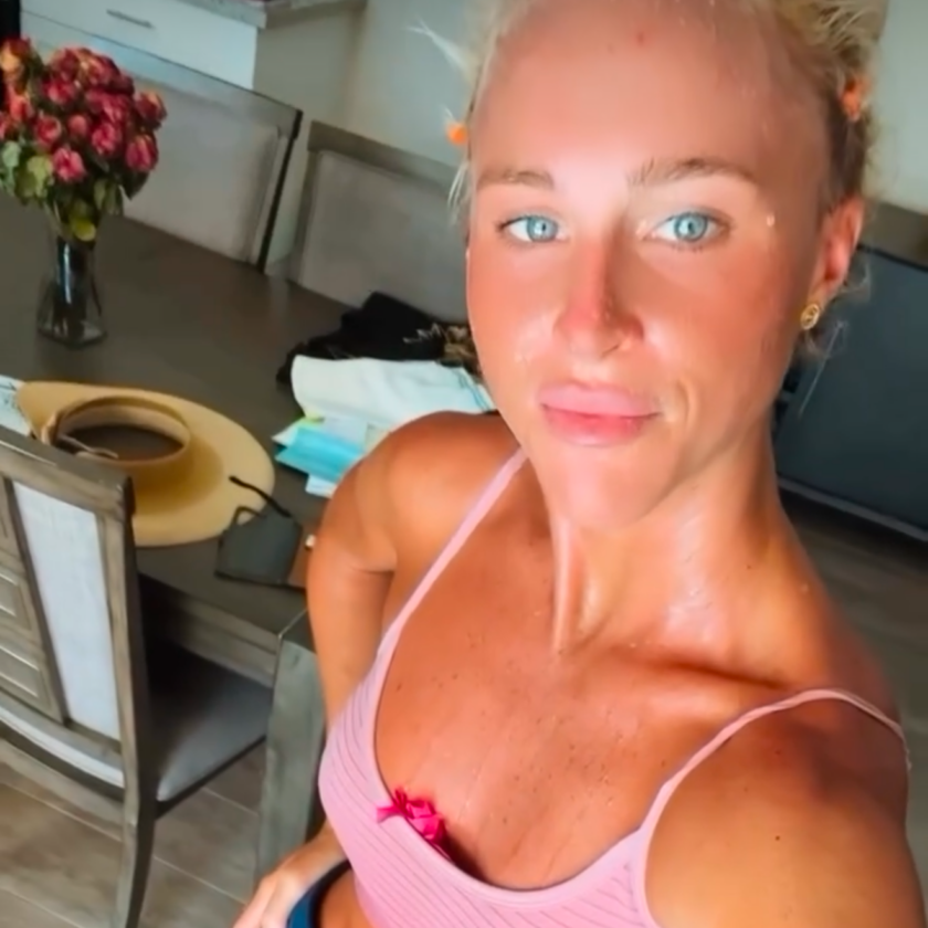 Selfie of a sunburned woman in a pink striped bikini top standing in a kitchen with a hat on a chair and a vase of pink flowers in the background