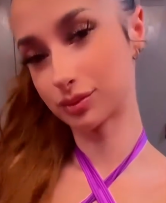 Young woman with long red hair wearing a purple cross-strap top, posing in a locker room with gray lockers behind her.