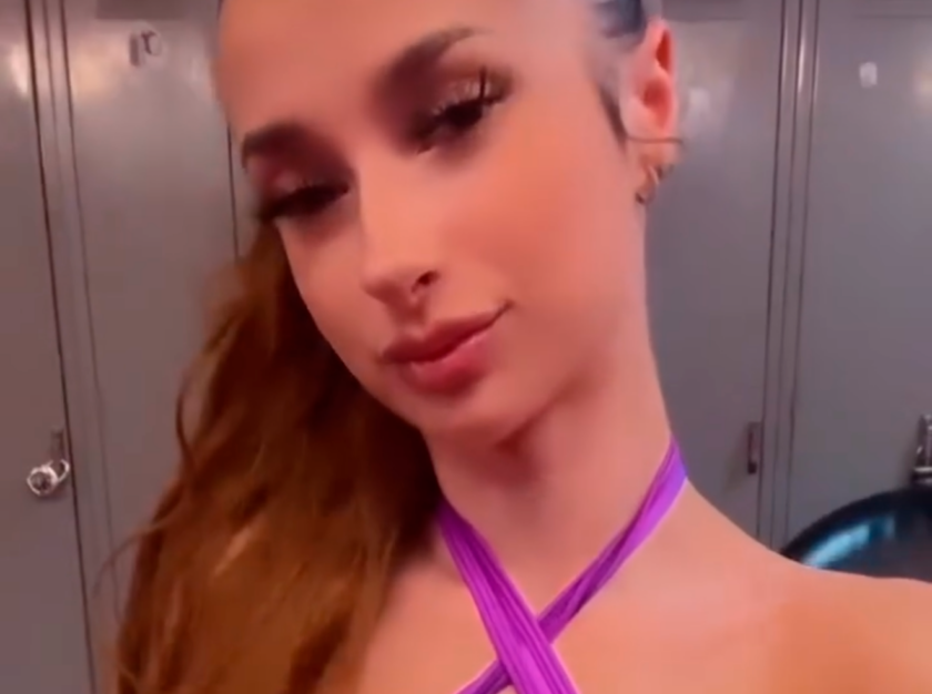 Young woman with long red hair wearing a purple cross strap top posing in a locker room with gray lockers behind her