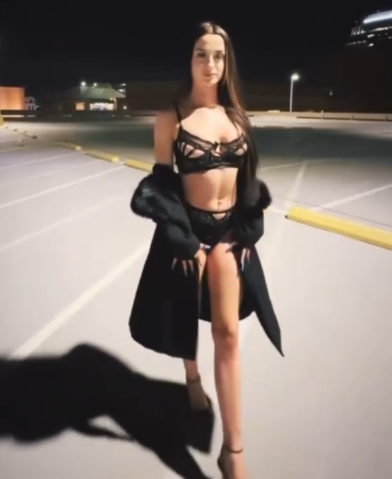 Woman in black lace lingerie and heels posing in a dimly lit parking lot at night with a coat draped over her arms