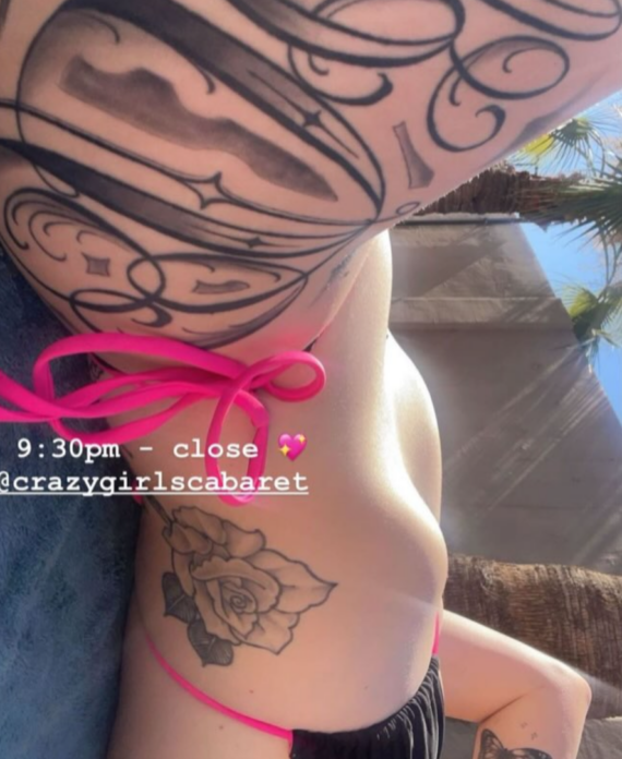 Person wearing a pink bikini bottom side view outdoors showing a large arm tattoo sleeve a rose on the side and butterfly tattoos on the leg
