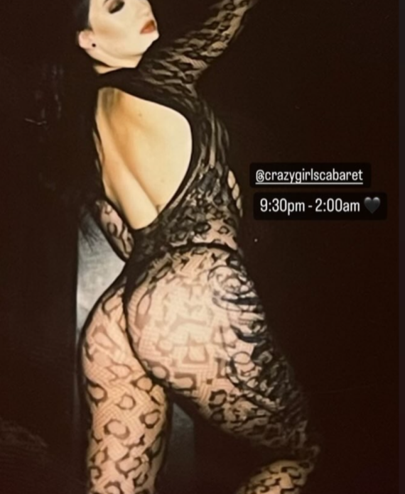 Woman in a black lace bodysuit striking a sultry side pose against a dark background with one hand resting on her head