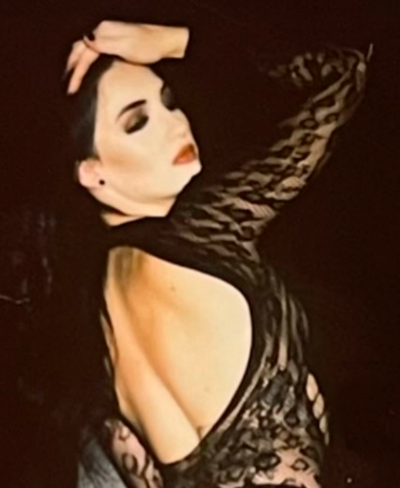 Glamorous portrait of a woman in a black lace dress, pale makeup, dark hair, posed with one arm raised over her head.