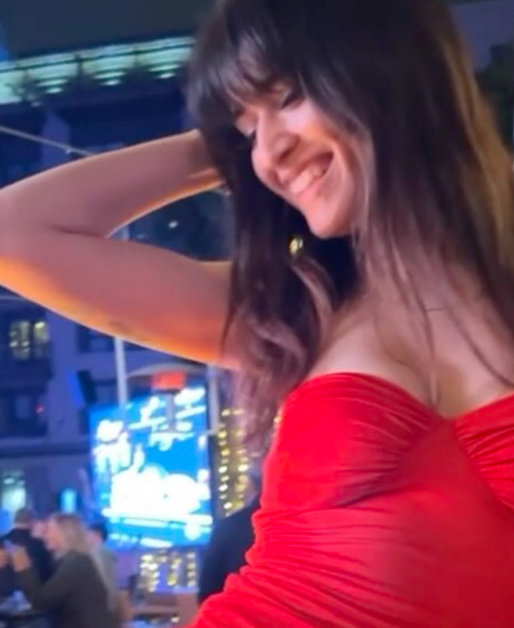 Woman in a red strapless dress smiles with one arm behind her head at a nighttime outdoor party with blue lights in the background.