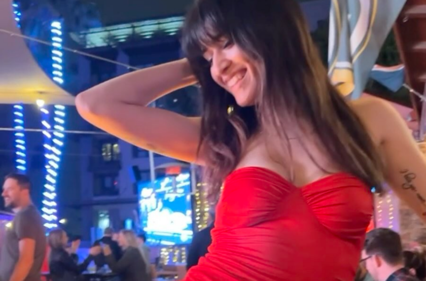 Woman in a red strapless dress smiles with one arm behind her head at a nighttime outdoor party with blue lights in the background
