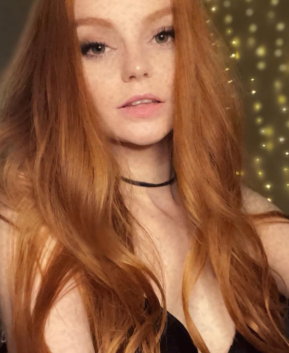 Portrait of a young woman with long red hair wearing a black top and a thin black choker looking at the camera with soft warm lighting