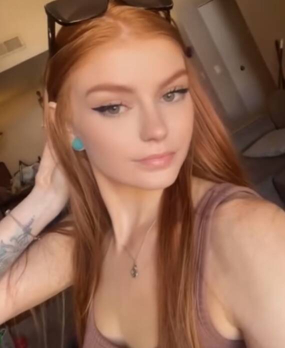 Young woman with long red hair taking a selfie indoors wearing a pink tank top and sunglasses on her head